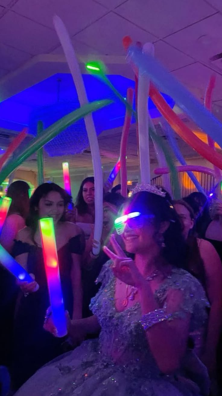 Quinceañera con lentes LED, corona y globos largos rodeada de invitados